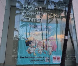 Glass exterior of the Hawaii Convention Center with a large “Kawaii Kon” banner inside. The banner features cute chibi characters, event dates for 2025. Photo by Ann Li.
